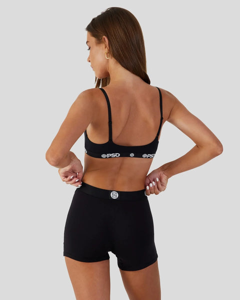 Woman wearing black sports bra with thin straps and matching black shorts with PSD branding, shown from back view.