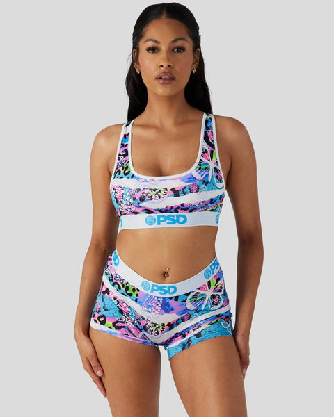 Woman wearing colorful neon floral print sports bra and matching boy shorts with white trim and blue branding.