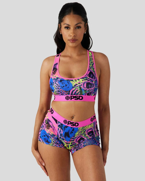 Woman wearing neon multicolored grid-patterned sports bra and boy shorts set with pink trim and PSD branding.