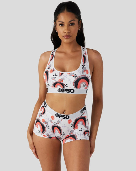 Woman wearing white sports bra and boy shorts with red and black abstract pattern and PSD branding.
