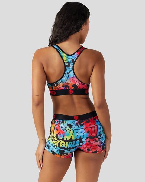 Woman wearing colorful Powerpuff Girls sports bra and shorts with blue, red, and yellow cartoon character print on neutral ba