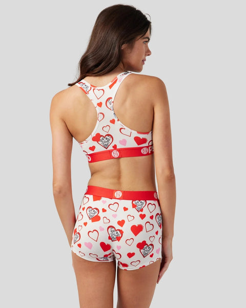 Woman wearing white racerback sports bra and matching shorts with red hearts and character print, red trim bands.