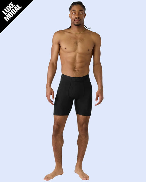 Man wearing black fitted shorts with fly detail, standing shirtless against light blue background.