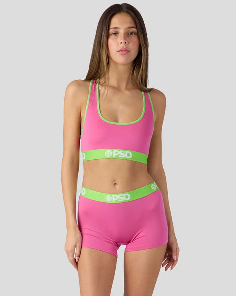 Woman wearing bright pink sports bra and matching shorts with green trim and PSD branding.