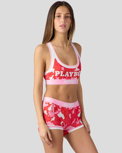 Woman wearing red and pink camouflage sports bra and shorts set with Playboy branding and white trim.