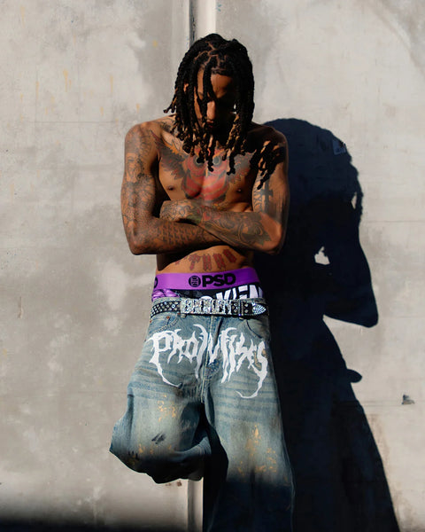 Shirtless man with dreadlocks and tattoos wearing purple waistband, studded belt, and distressed denim jeans against white wa