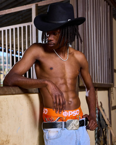 Man wearing black cowboy hat, silver chain necklace, orange PSD waistband, and light blue jeans in barn setting.