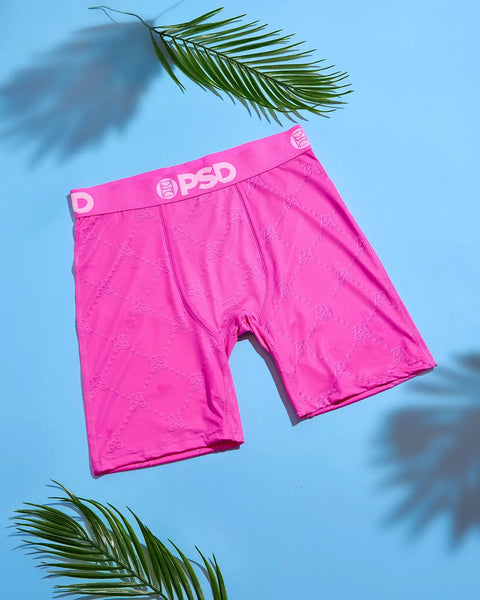 Bright pink boxer briefs with monogram pattern and PSD waistband displayed on blue background with palm fronds.
