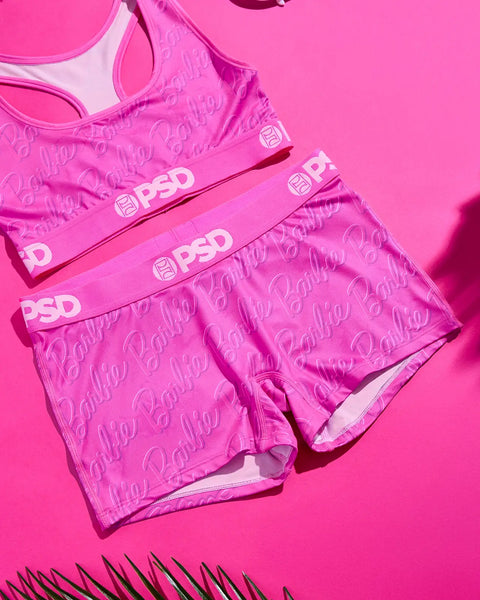 Hot pink sports bra and boy shorts set with "Barbie" all-over print and PSD waistband against pink background.