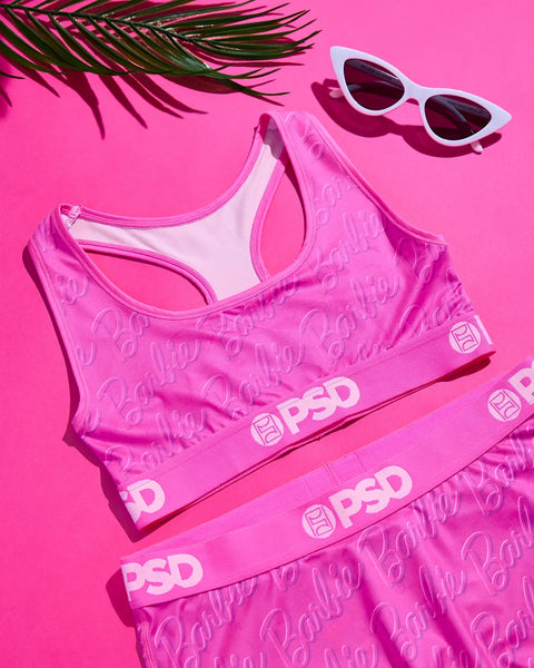 Pink sports bra and underwear with Barbie branding, white cat-eye sunglasses, and palm frond on hot pink background.