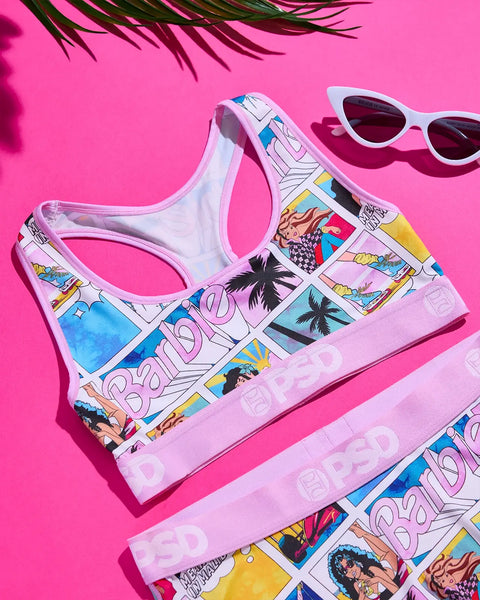White sports bra with colorful comic-style Barbie graphics, displayed with white cat-eye sunglasses on bright pink background