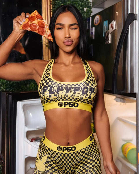 Woman wearing yellow and black checkered Playboy sports bra and matching briefs holding pizza slice.