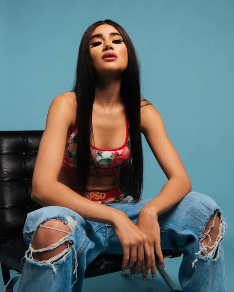 Woman wearing red floral crop top with ripped blue jeans, seated against blue background.