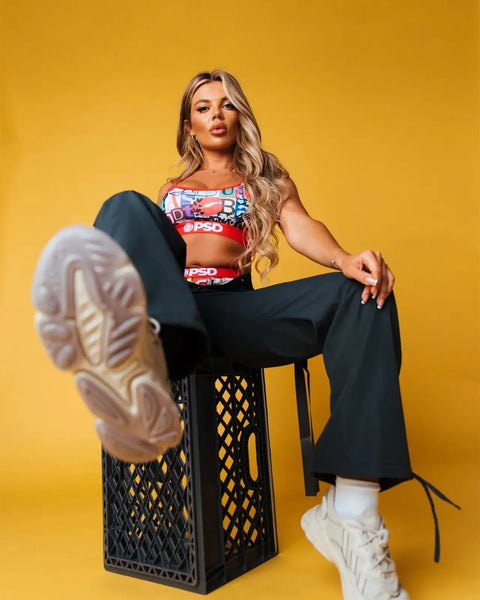 Woman wearing red and white patterned sports bra with black pants and cream sneakers, seated against yellow background.