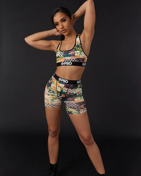 Woman wearing multicolor patchwork sports bra and matching shorts with checkered and camouflage pattern, black waistband bran