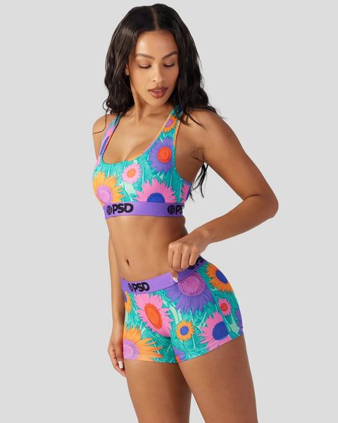 Woman wearing colorful multiflower-print sports bra and matching shorts with purple waistbands against white background.