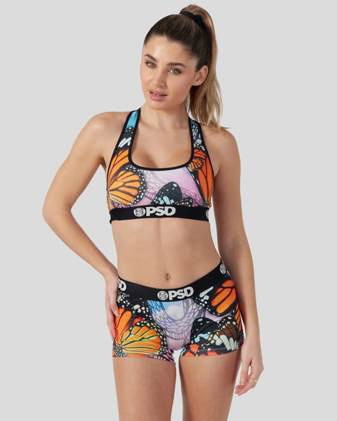 Woman wearing sports bra and shorts set with orange and black butterfly print and PSD branding.