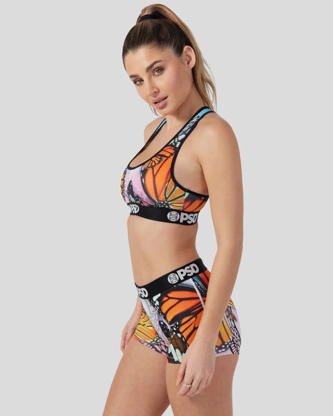 Woman wearing sports bra and shorts set with orange and purple butterfly wing print and black PSD branding.