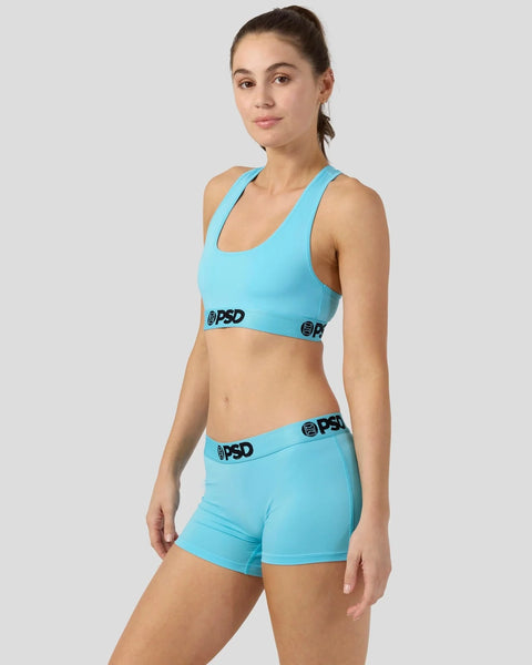 Woman wearing bright turquoise racerback sports bra and matching boy shorts with PSD branding.