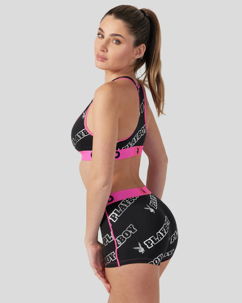 Woman wearing black and hot pink sports bra and shorts set with Playboy branding and repeating text pattern.
