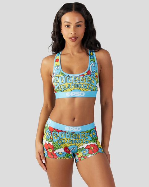 Woman wearing colorful Cali Cookies-themed sports bra and shorts set with floral and text print on light blue background.