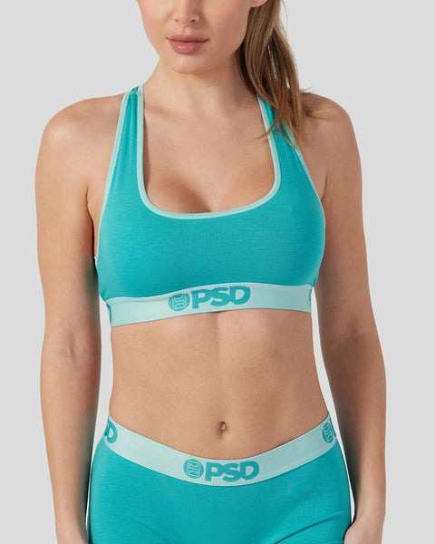 Woman wearing a jade green sports bra with light blue trim and PSD branding.