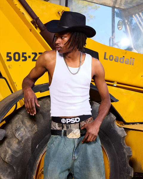 Man wearing black cowboy hat, white tank top, and camouflage-patterned belt sitting on yellow heavy machinery tire.