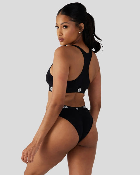 Woman wearing black racerback sports bra and high-waisted bikini bottoms with logo details, shown from back angle.