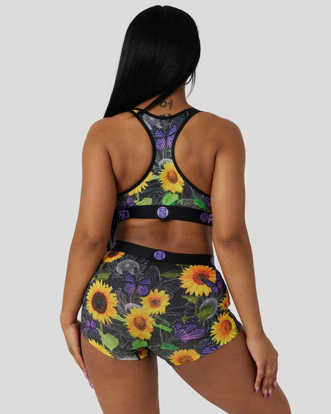 Sports bra and shorts set with sunflower and butterfly print on dark background, black straps and purple snap details.