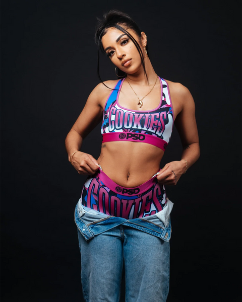 Woman wearing pink, blue, and white Cookies brand sports bra and matching waistband with camo pattern over blue jeans.