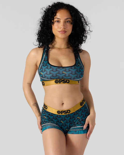 Teal sports bra and shorts set with geometric pattern and gold-trimmed waistband, worn by model with black curly hair.