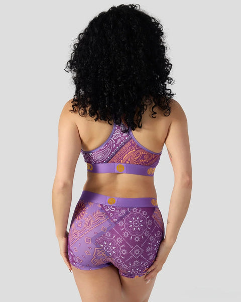 Purple bandana-print two-piece swimsuit with paisley and geometric patterns, shown from back view on model with black curly h