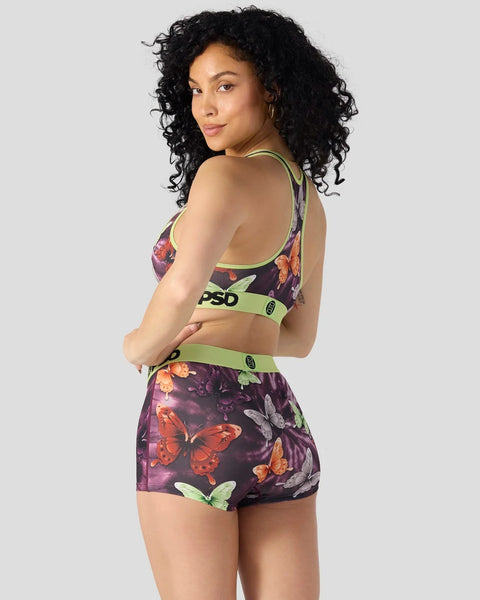 Woman wearing dark purple sports bra and shorts set with multicolored butterfly print and neon green bands.
