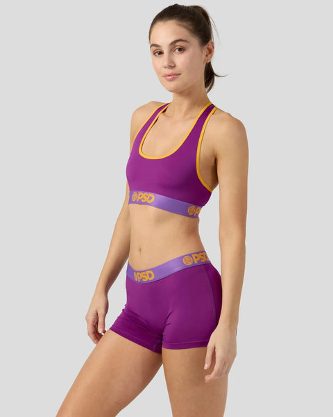 Woman wearing purple sports bra and matching shorts with yellow trim and logo detailing.