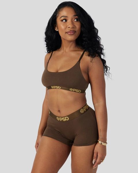Woman wearing a cocoa-brown sports bra and matching shorts set with gold branded waistband trim.