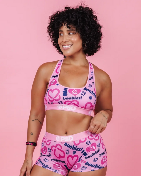 Woman wearing pink sports bra and shorts set with heart and text print against pink background.