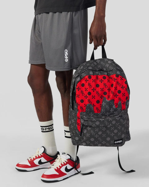 Black backpack with red and gray patterned front panel, black shoulder straps, held by person wearing gray shorts and red sne
