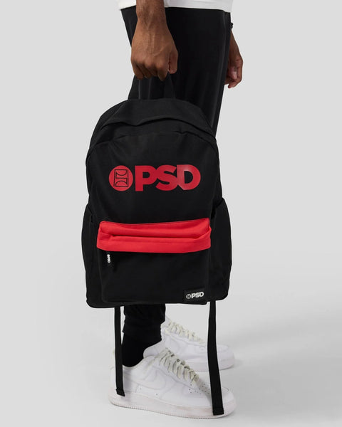 Black backpack with red PSD logo on back and red zippered pocket, worn by person in white sneakers.