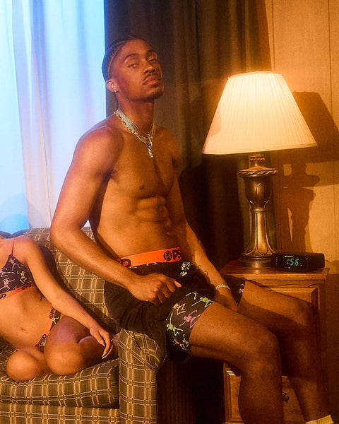 Shirtless man sitting on plaid couch wearing patterned shorts and chain necklace in warm-lit bedroom setting.