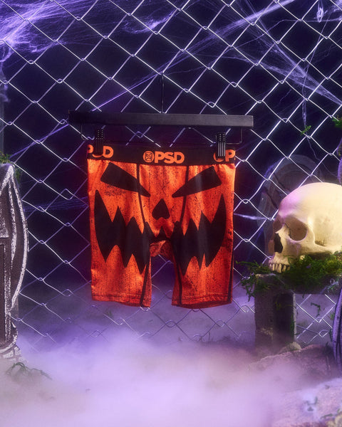 Red PSD boxer shorts with jack-o'-lantern face design displayed on hanger against purple neon grid backdrop.
