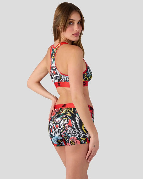 Woman wearing red racerback bikini top and matching shorts with skull and multicolor tattoo-style print throughout.