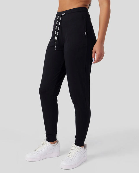 Black joggers with drawstring waist and white text branding, tapered ankles, paired with white sneakers.