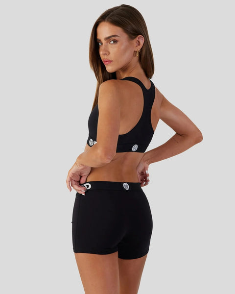 Woman wearing black sports bra and matching black shorts with logo details, shown from back angle.