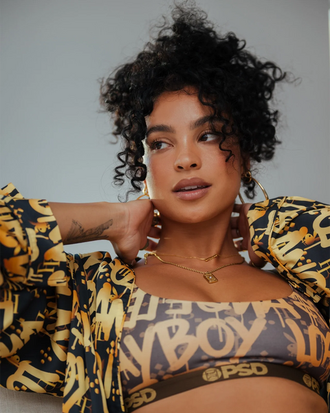 Woman wearing yellow and black graffiti-print Playboy bra top with matching oversized shirt and gold chain necklace.