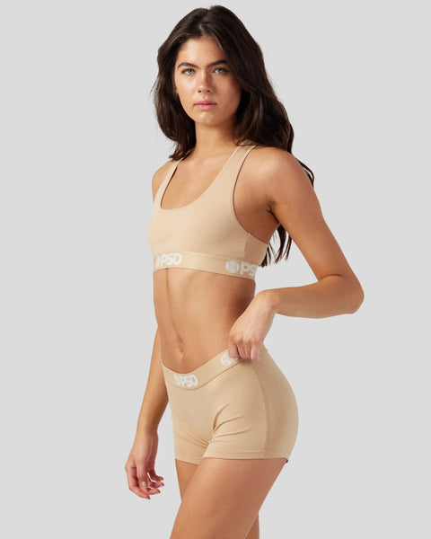 Woman wearing sand-colored sports bra and matching shorts with branded waistband detail.