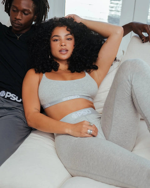 Woman wearing grey athletic crop top and matching leggings, seated on white couch.