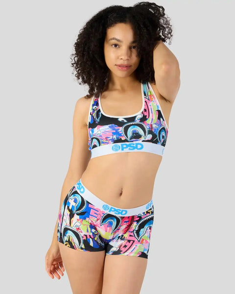 Woman wearing colorful shark-patterned sports bra and shorts with white PSD waistbands against gray background.