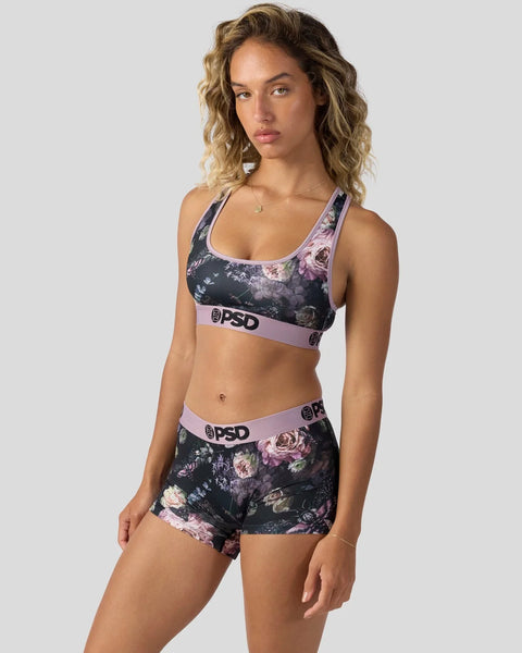 Woman wearing dark floral sports bra and shorts set with pink elastic waistbands and branding.