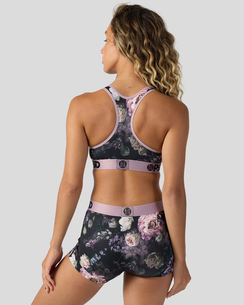 Woman wearing dark floral sports bra and shorts set with pink trim and buckle details, shown from back view.