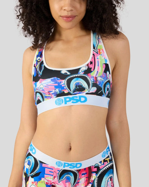 Woman wearing a PSD sports bra with colorful shark and geometric print on black background with white band.